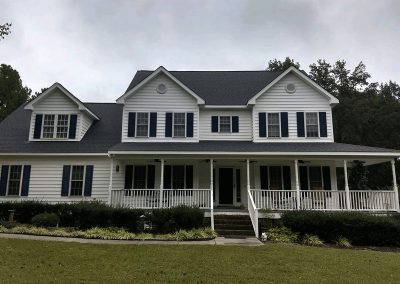 White two-story farmhouse with dormers, a wraparound porch, and a newly installed dark gray shingle roof.