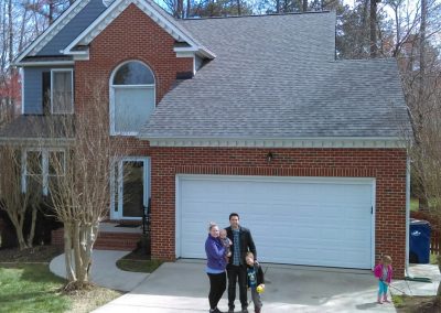Family standing in the driveway of their brick home after a new gray shingle roof installation.