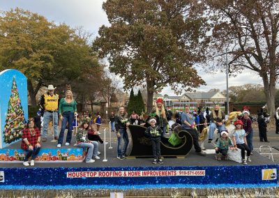 Families riding the Housetop Roofing & Home Improvements sleigh float beside a decorated Christmas tree display.