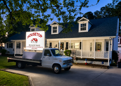 Housetop Roofing sign truck parked in front of a white dormered house at dusk.